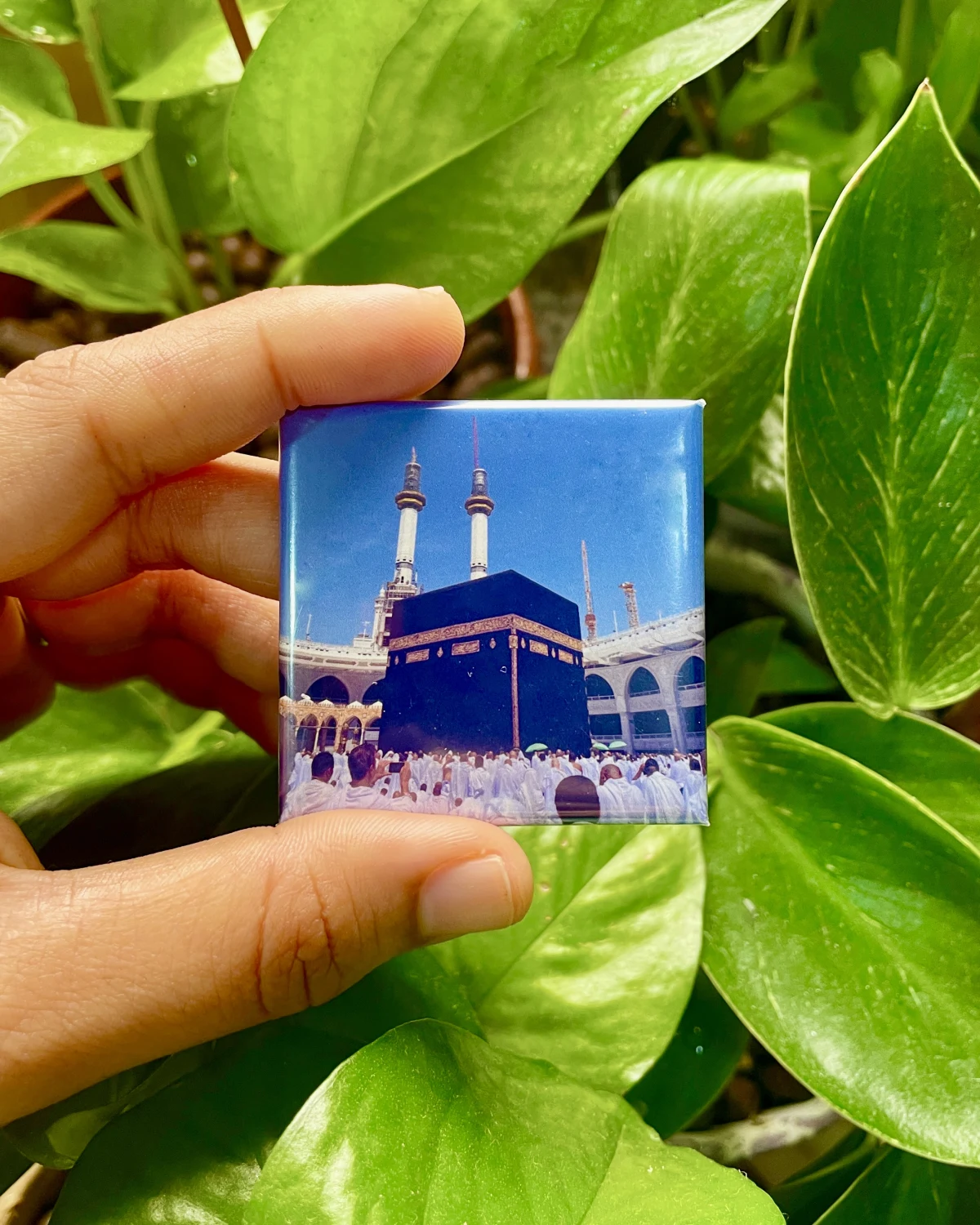 A classic 3D photo magnet of the Kaaba held between two fingers in front of green leaves